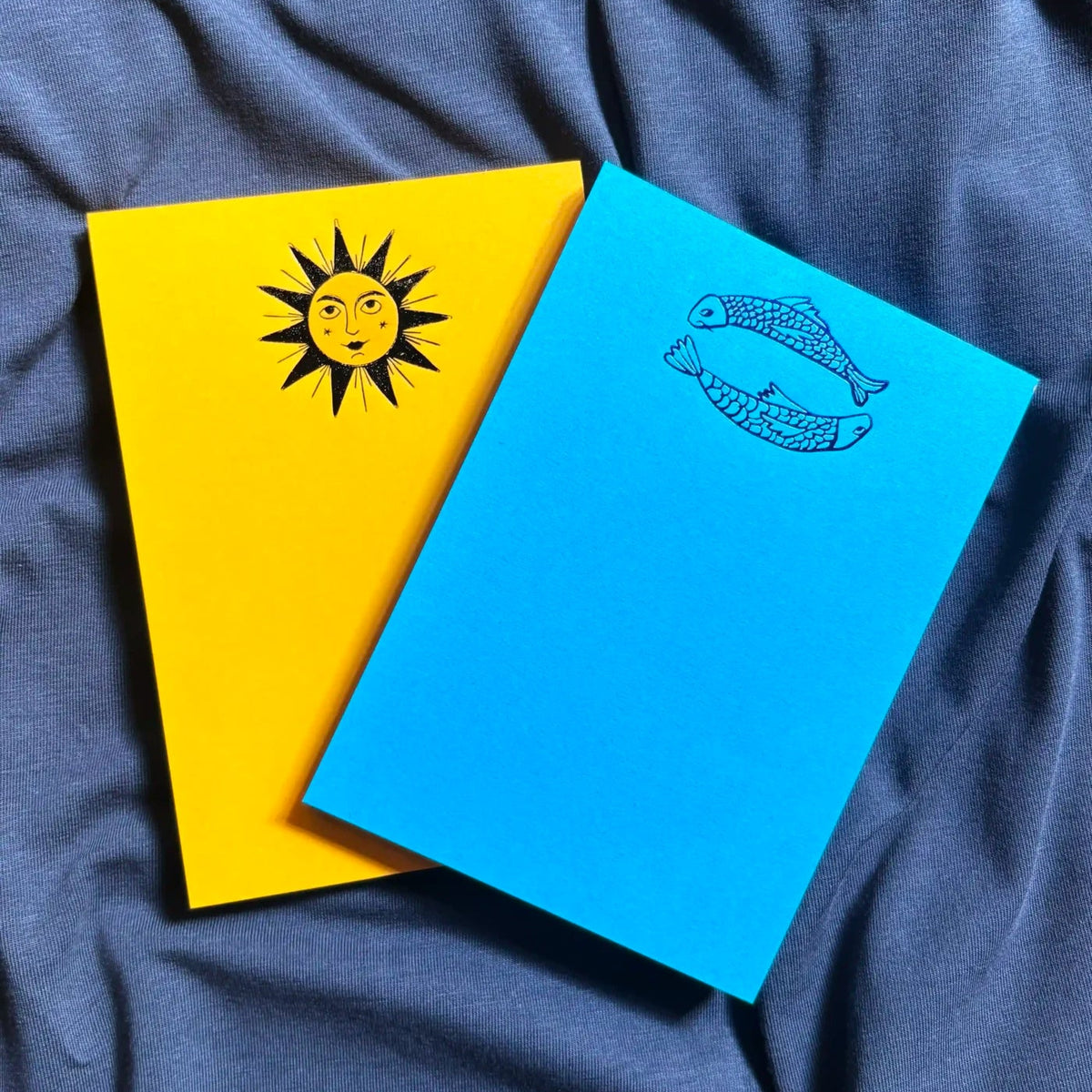 Two small notebooks or cards with bright yellow and blue covers, placed on a dark blue fabric background. The yellow cover has a black sun illustration, and the blue cover has a black fish illustration.