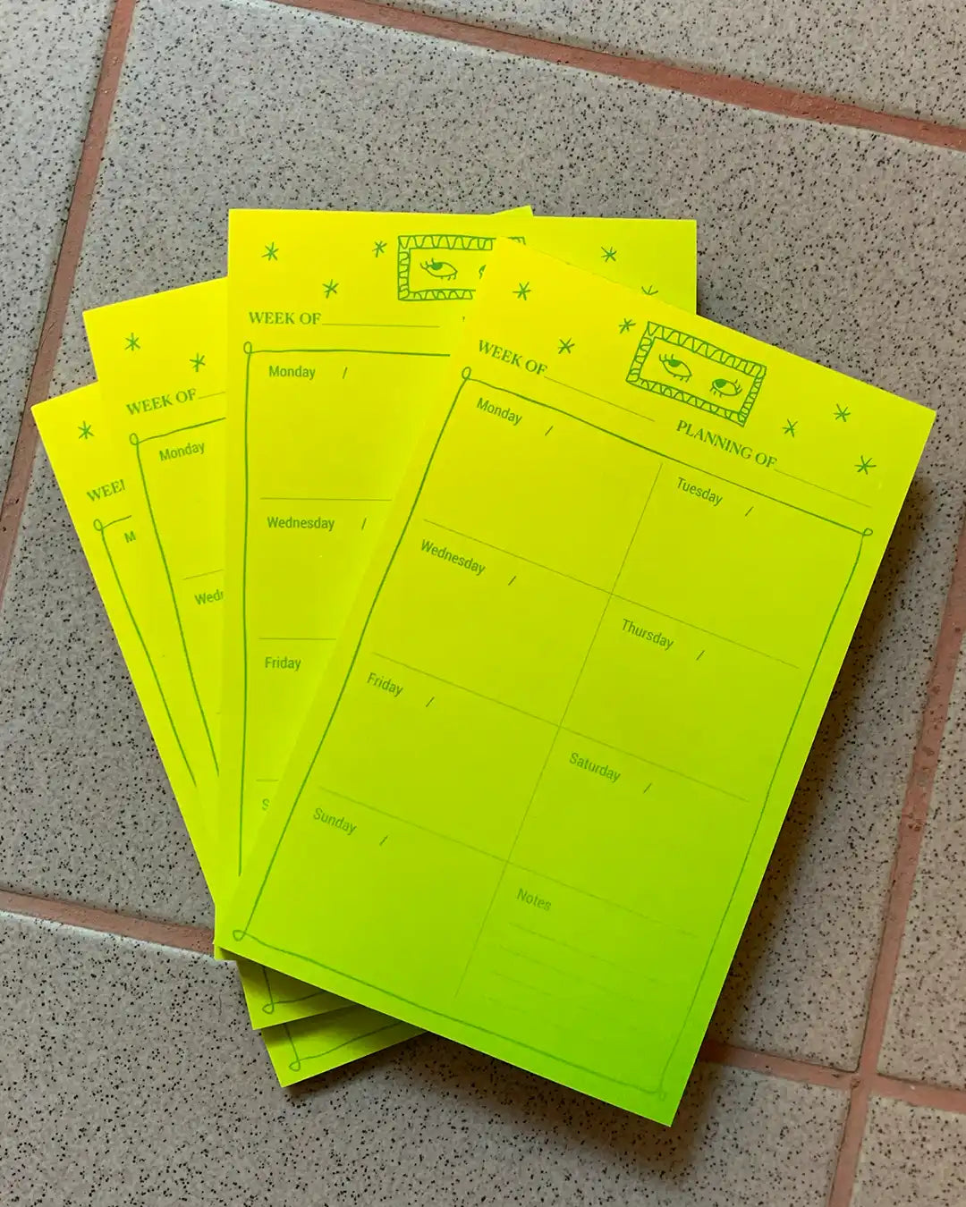 A stack of bright neon yellow weekly planning pads with green printed text and grid sections, placed on a speckled gray tiled floor.