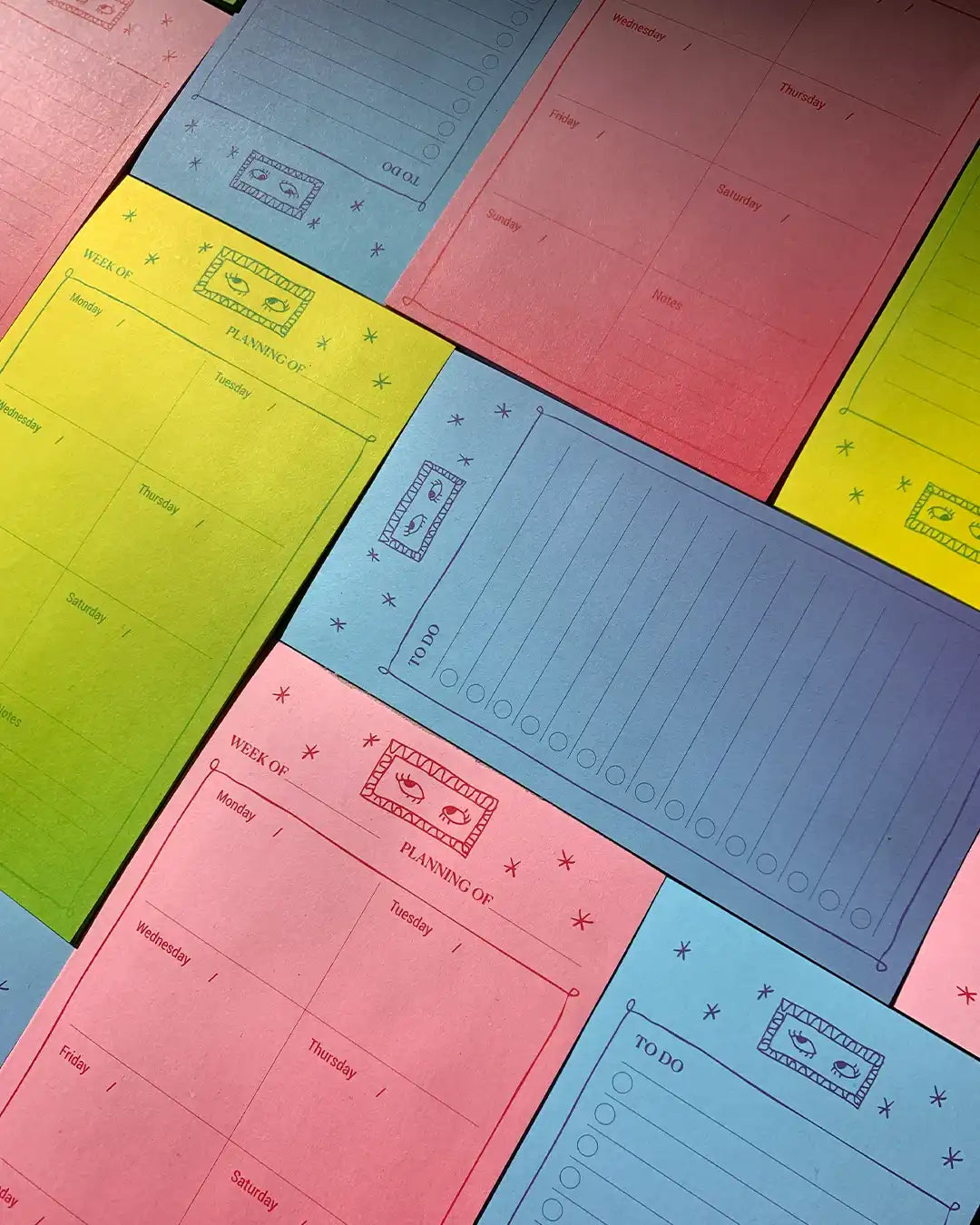 A close-up, angled view of several pastel-colored daily planning pads overlapping. The pads are in shades of pink, blue, yellow, and green, each printed with daily sections, a weekly planner grid, and a 'To Do' list.