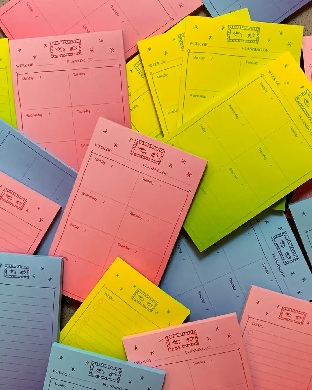 A pile of colorful weekly planning notepads in pink, yellow, blue, and purple, showing the front cover design with a cartoon character and the text 'WEEK OF PLANNING'.
