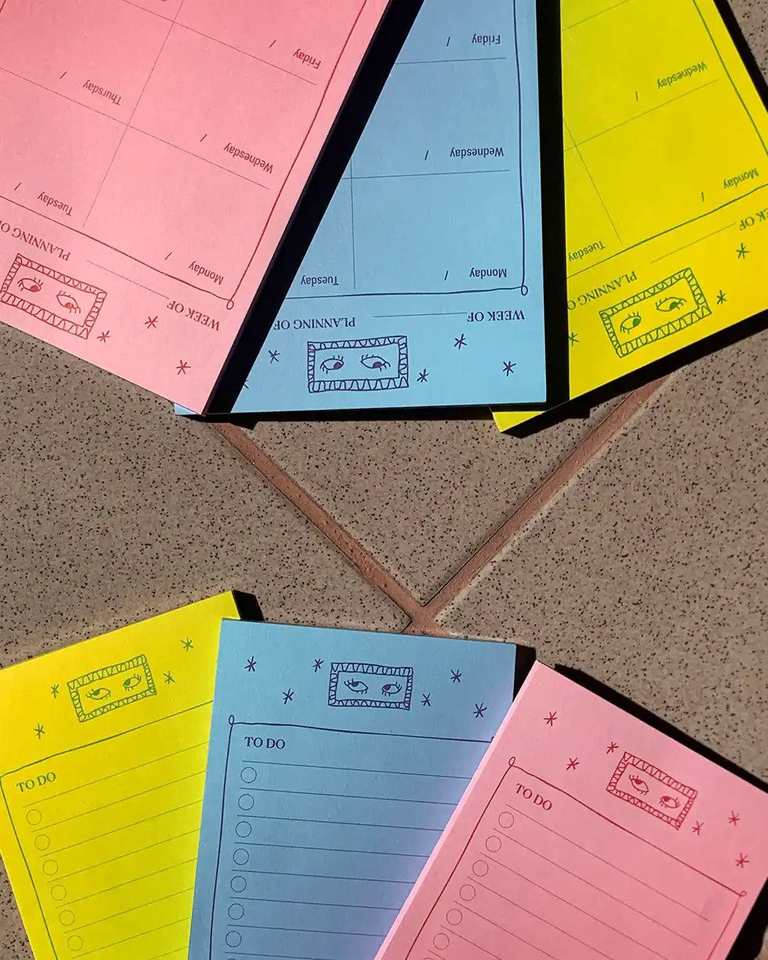Colorful weekly planner notepad sheets in pink, light blue, and yellow arranged on a speckled tile surface. The notepads feature sections for Monday through Sunday, a weekly summary box, and a to-do list.