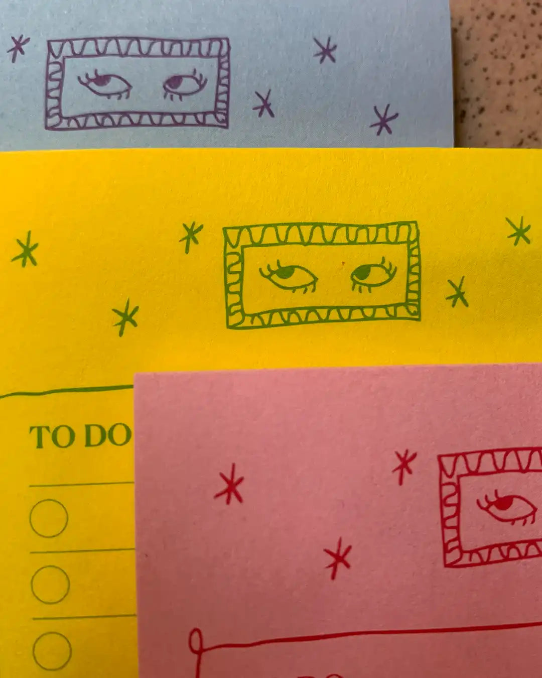 Three colorful sticky notes or paper cards stacked together, each printed with a hand-drawn eye inside a rectangular frame, plus small star-like marks. The top note is light blue with purple drawing, the middle is yellow with green drawing, and the bottom is pink with red drawing.