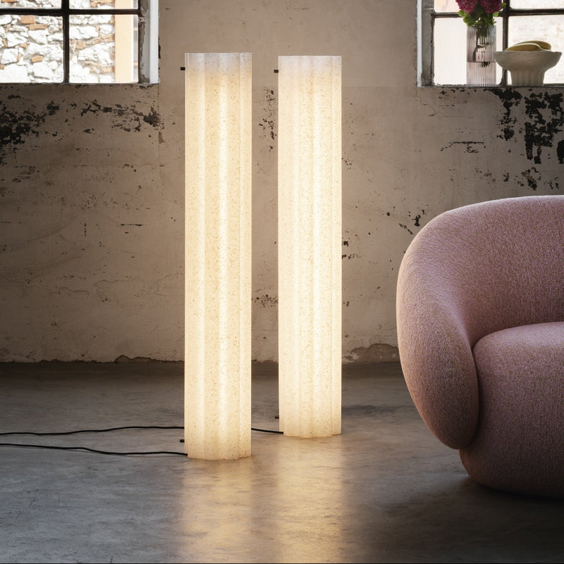 Two tall cylindrical floor lamps with a textured off-white fabric, standing in a rustic room with a concrete floor, textured plaster walls, and a pink armchair partially visible on the right.