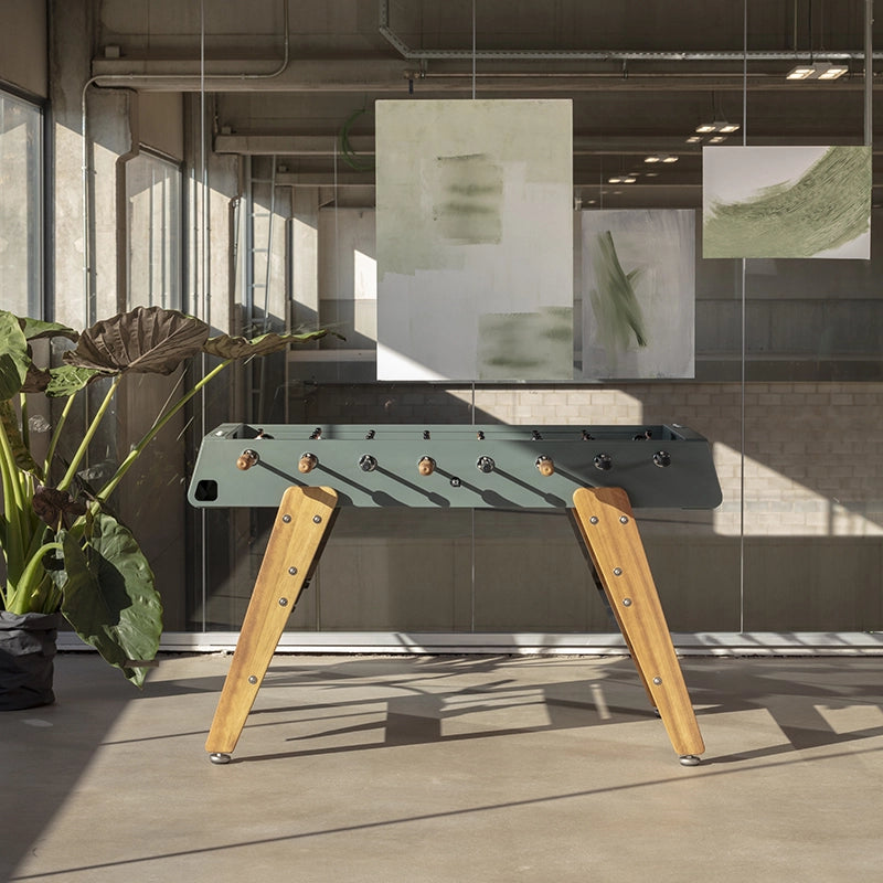 A modern foosball table with a dark green top and light wood legs, positioned in a contemporary loft-style room with large windows, plants, and abstract wall art.